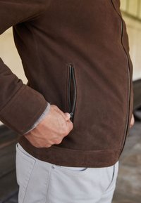 Chaqueta de ante marrón con un bolsillo lateral con cremallera. La tela tiene una textura suave, y la chaqueta presenta un diseño entallado y puños acanalados.