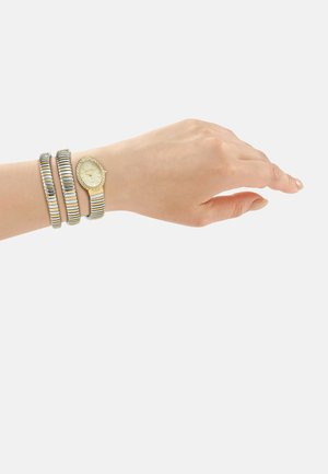 Pols met goud- en zilverkleurige gestreepte wikkelhorloge met een ovalen wijzerplaat en een gestructureerde lunet tegen een effen lichte achtergrond.