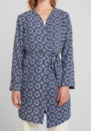 Blå mønstret slå-om-frakke med cirkulære designs, lange ærmer og taljebånd, båret over hvide bukser.