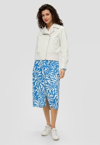Witte ritsjack met een cropped ontwerp, gecombineerd met een blauwe rok met een wit abstract patroon. Model draagt witte sneakers.