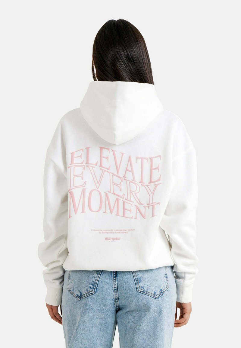 Felpa bianca con testo rosa "ELEVATE EVERY MOMENT" sulla schiena. Tessuto morbido, cappuccio spazioso e polsini a coste. Abbinata a jeans azzurri chiari.