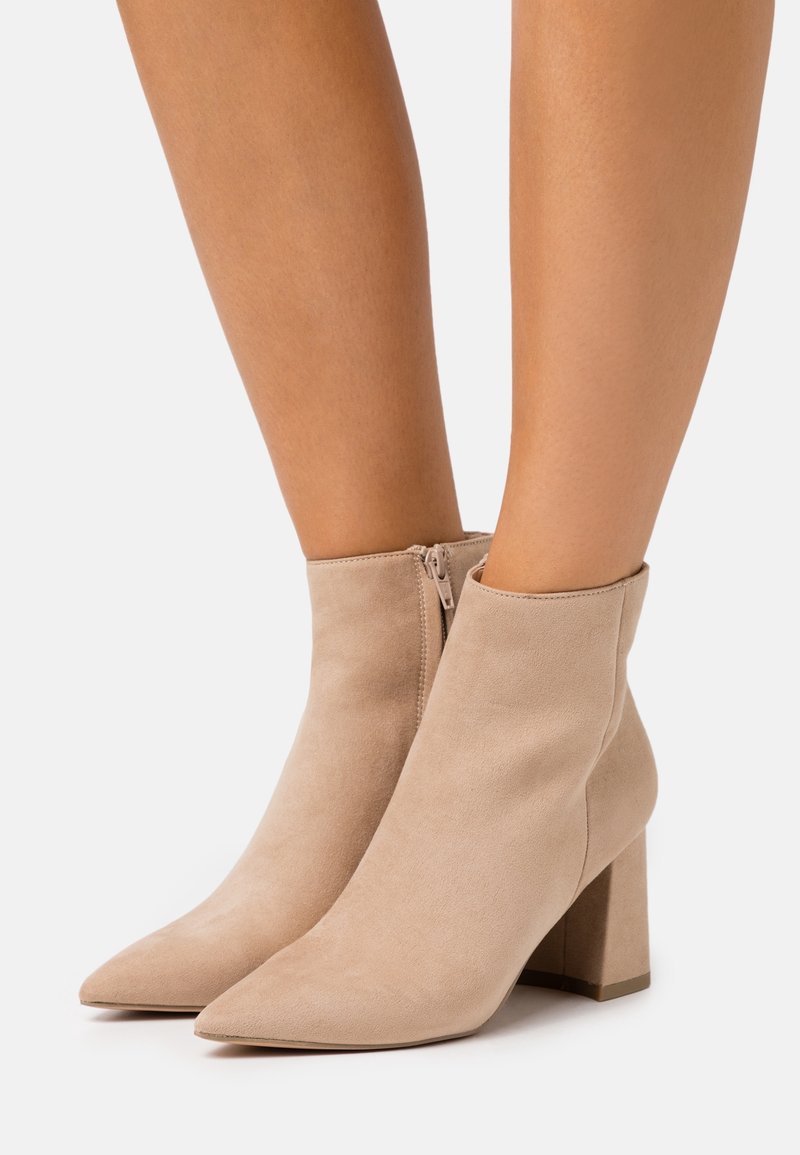 Femme portant des bottines en daim beige à bouts pointus et talons blocs, debout sur un fond blanc.