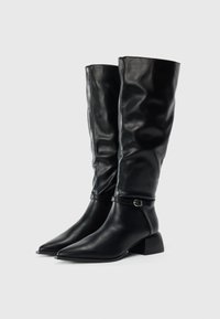 Par de botas de cuero negro hasta la rodilla con puntas afiladas, tacones bajos y un delgado cinturón con hebilla alrededor del tobillo.