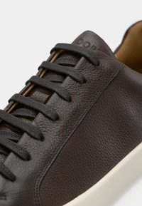 Brun lädersneaker med en texturerad yta, svarta snören och en krämfärgad sula. Bruna detaljer framhäver designen.