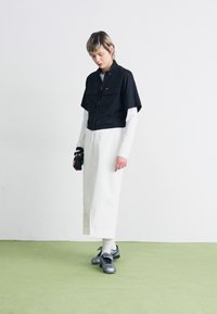 T-shirt noir court avec deux poches sur la poitrine, superposé à une longue robe blanche. Des baskets grises et des chaussettes blanches complètent la tenue. Sol en moquette verte.