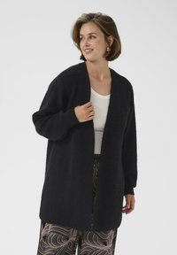 Femeie îmbrăcată cu un cardigan negru cu deschidere frontală, un top alb și pantaloni largi cu model, zâmbind și privind în lateral pe un fundal simplu.
