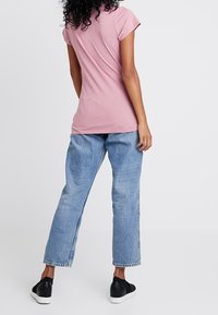 Růžové bavlněné tričko s krátkým rukávem, spárované se světle modrými džíny. Model má na sobě černé slip-on boty, pohled zezadu.