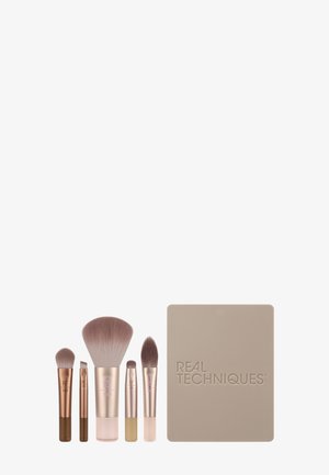 Five makeup brushes with rose gold handles arranged beside a beige rectangular Real Techniques brush holder.