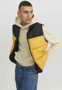 Chaleco tipo plumas con paneles negros y amarillos, con cremallera frontal, cuello alzado y textura acolchada, usado sobre una sudadera beige.