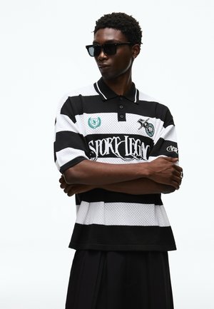 Homme portant des lunettes de soleil noires et un polo rayé noir et blanc avec texte graphique et écussons, bras croisés, regardant sur le côté.