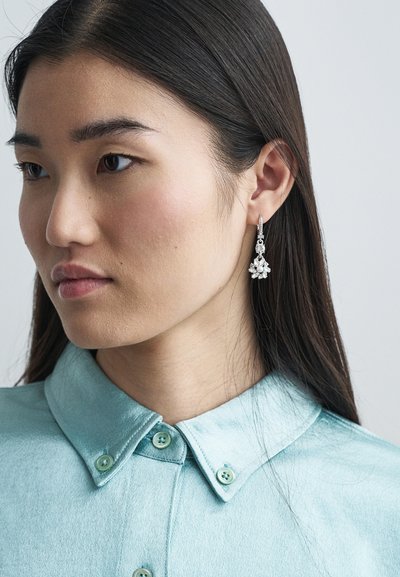 Mlada ženska z dolgimi temnimi lasmi, oblečena v svetlo modro srajco s kratkimi rokavi in okrasnimi srebrnimi uhančki s kristali, gleda levo.