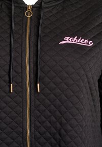Sort quiltet hættejakke med lynlås og lyserød "achicoc" logo, guldfarvet lynlåsdæksel og sorte snore. Har en tekstureret overflade og sporty design.