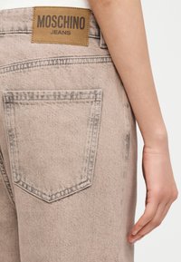 Πίσω όψη ανοιχτό μπεζ τζιν με ετικέτα μάρκας Moschino σε καφέ χρώμα στη μέση και το δεξί χέρι ενός ατόμου δίπλα του.