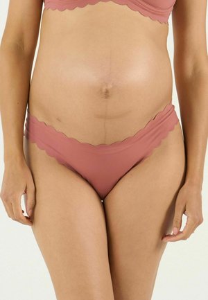 Frauenmitte, die ein passendes Unterwäsche-Set in Mauve mit gewelltem Rand trägt, vor einem schlichten hellen Hintergrund stehend.