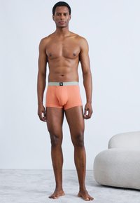 Cuecas boxer laranja com cintura cinza clara, feitas de tecido liso. Design ajustado que delineia o tronco e as pernas.