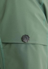 Green fabric shirt featuring a black button detail with a raised emblem. Smooth texture, streamlined design, and contrasting stitching visible.
