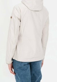 Personne portant une veste à capuche beige clair à manches longues et un jean bleu, vue de dos sur un fond blanc.