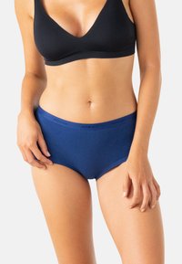 Femme portant un soutien-gorge noir et un slip taille haute bleu avec une impression de marque subtile sur la ceinture, se tenant devant un fond clair.