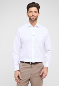 Weißes Langarmhemd mit Knopfkragen, Tasche auf der linken Brust und silbernen Knöpfen, kombiniert mit hellbraunen Hosen.