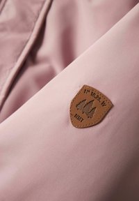 Svijetloružičasta tkanina s glatkom teksturom i smešnom kožnom oznakom koja prikazuje logotip drveta i tekst, prišivena na jaknu.