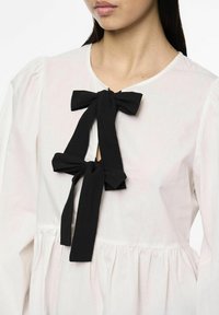 Blouse blanche à encolure ronde, avec un détail en forme de clé noué par deux nœuds en tissu noir. Bas avec volants et manches bouffantes.