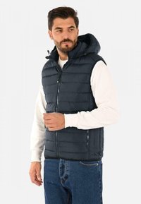 Gilet imbottito blu navy con cappuccio, chiusura frontale con zip e tasche laterali. Presenta un design trapuntato e una texture liscia, indossato sopra una shirt bianca.