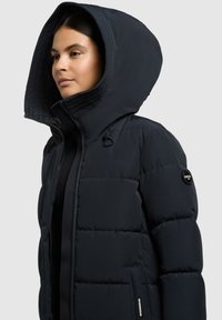 Chaqueta de plumas en azul marino oscuro, con una gran capucha, textura acolchada, cierre de cremallera y detalle de logo en la manga.