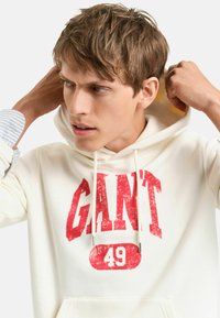 Kremowa bluza z kapturem z czerwonym, przetartym nadrukiem "GANT 49", z sznurkami do regulacji i miękką fakturą. Model dostosowuje kaptur, ukazując warstwową koszulę pod spodem.