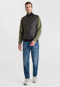 Gilet imbottito nero con design trapuntato sopra una felpa verde grigio, abbinato a jeans cargo blu e sneaker chiare. Modello maschile in piedi.