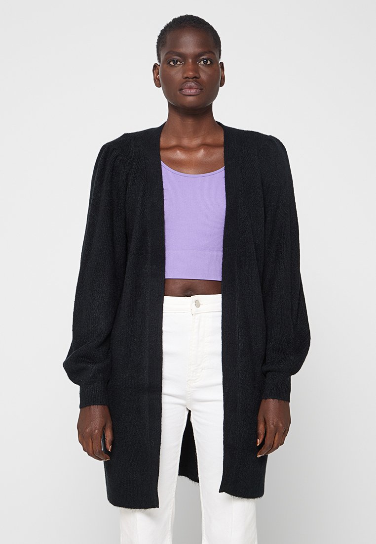 Zwarte lange cardigan van textuurstof, met open voorkant en pofmouwen, gedragen over een lavendelkleurige crop top en witte broek.