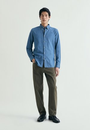 Hombre de pie contra un fondo liso, vestido con camisa azul abotonada, pantalones verde oliva y zapatos negros, con una mano en el bolsillo.