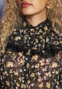 Personai ar lokainiem matiem, kas valkā melnu ziedu blūzi ar pērļu pogām un mežģīņu volānu apkakli, lūpas nedaudz atvērtas un neitrāls fons.