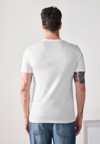 T-shirt en coton blanc à manches courtes et col rond, avec une texture lisse. L'individu a un tatouage visible sur le bras.