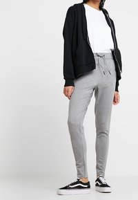 Černá mikina na zip, bílý tričko, šedé tepláky a černé tenisky. Materiál je měkký; kalhoty mají elastický pas se šňůrkou.