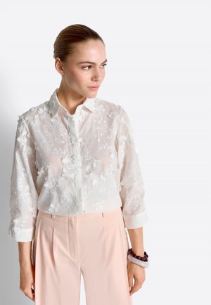 Kobieta z włosami związanymi do tyłu, ubrana w białą prześwitującą bluzkę z haftem kwiatowym oraz jasnoróżowe spodnie, patrząca w lewą stronę.