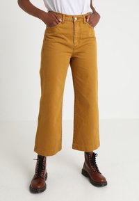 Pantalones de pierna ancha color mostaza hechos de algodón, con una cintura alta y bolsillos frontales, combinados con botas de tobillo de encaje marrones.