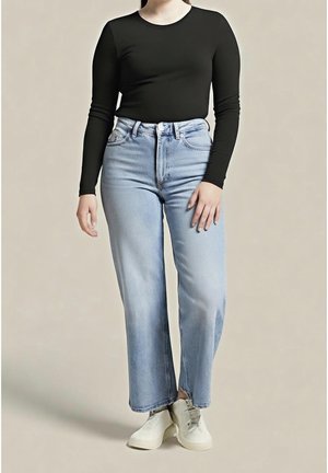 Personne portant un t-shirt noir à manches longues, un jean droit taille haute bleu clair et des baskets blanches, debout devant un fond uni.