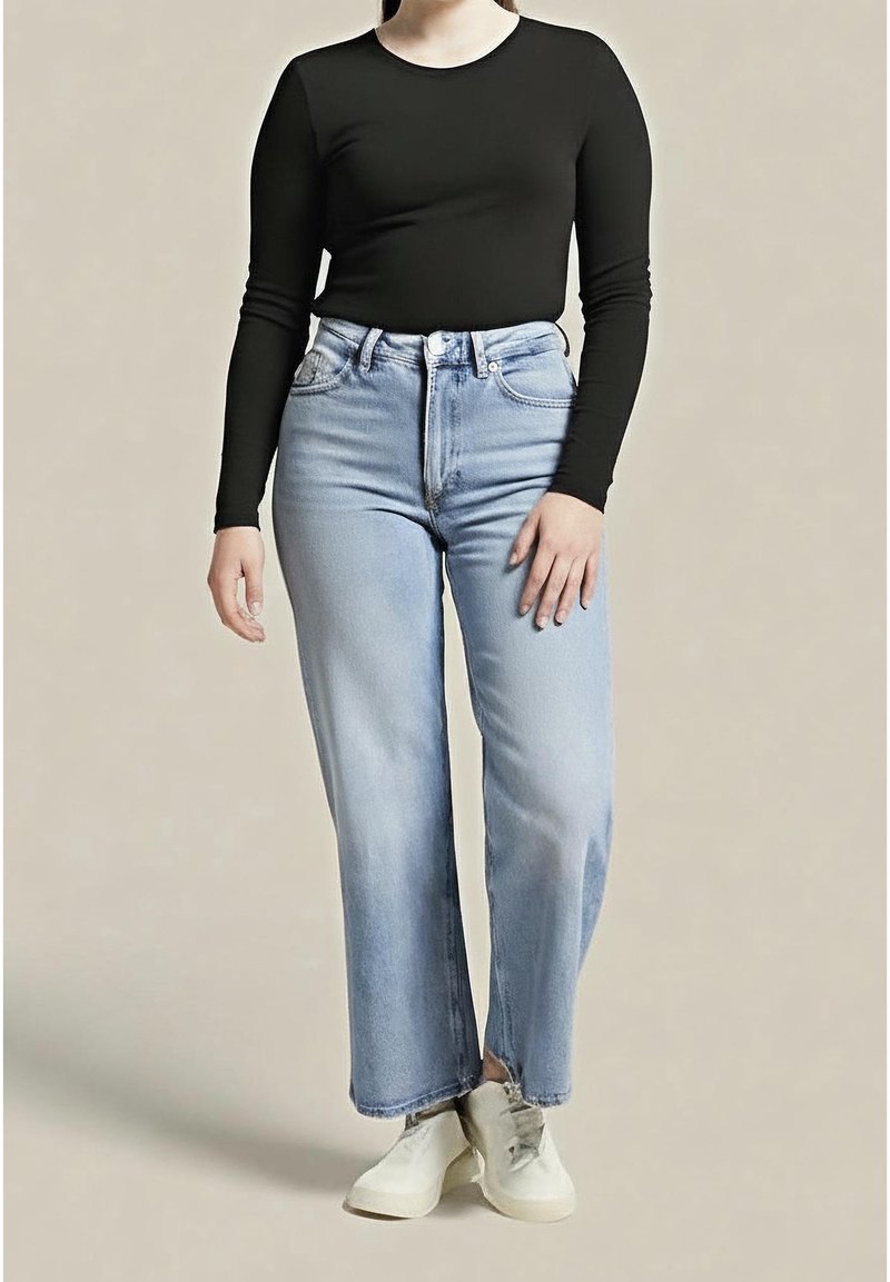 Personne portant un t-shirt noir à manches longues, un jean droit taille haute bleu clair et des baskets blanches, debout devant un fond uni.