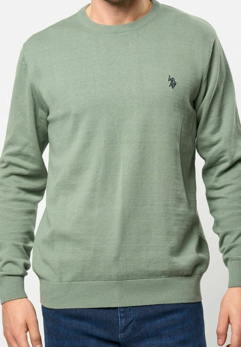 Homme portant un pull col ras-du-cou vert sauge avec un petit logo noir brodé sur le côté gauche de la poitrine, assorti à un jean bleu foncé.
