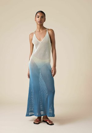 Žena v modro-bílé ombré síťované maxi šaty bez rukávů a červených žabek stojí před béžovým pozadím.