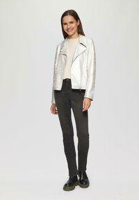 Giacca da moto in argento metallico con cerniera diagonale, indossata sopra una maglietta beige chiaro, abbinata a jeans skinny neri e stivali neri chunky.