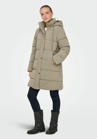 Beige polstret frakke med hætte, der har lynlås, to sidelommer og elastiske snore. Bæres over mørke jeans og sorte støvler.