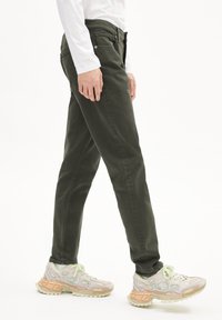 Khaki groene jeans met een aansluitend ontwerp, voorzien van vijf zakken en een gladde textuur. Gecombineerd met lichtgekleurde, gestructureerde sneakers.