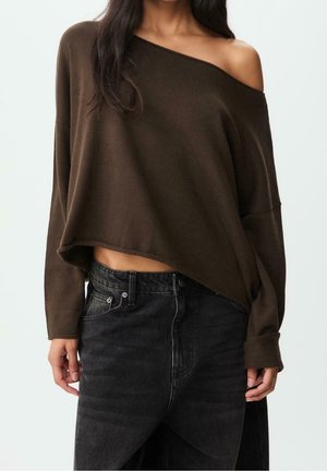 Femme portant un pull marron surdimensionné tombant sur une épaule et un jean en denim noir taille haute, coupé à mi-torse, debout devant un fond uni.
