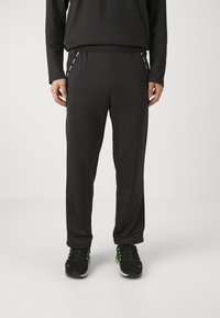 Schwarze Sporthose mit elastischem Bund, seitlichen Streifen und einem glatten, leichten Stoff. Getragen mit schwarzen Turnschuhen, die mit Grün akzentuiert sind.
