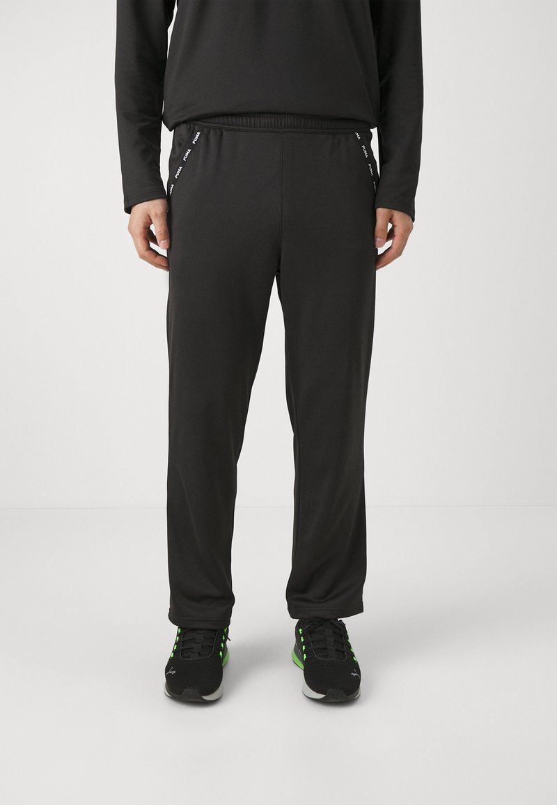 Schwarze Sporthose mit elastischem Bund, seitlichen Streifen und einem glatten, leichten Stoff. Getragen mit schwarzen Turnschuhen, die mit Grün akzentuiert sind.