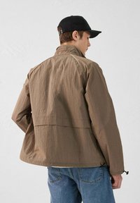 Bruine textuurjack met een hoge kraag, verstelbare taillekoord en raglanmouwen, gecombineerd met blauwe denim jeans.