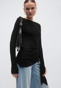 Bluză neagră cu mâneci lungi, cu un design răsucit în talie, asortată cu blugi evazați deschiși la culoare și o geantă neagră de umăr. Material moale, croială mulată.