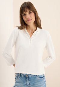 Witte long sleeve polo met kraag en drie knopen, gecombineerd met lichtblauwe jeans met zakken en een knoopsluiting.