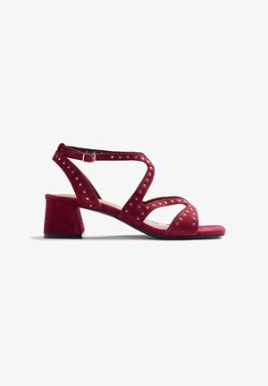 Sandale à talon en daim rouge avec bride à la cheville, bout ouvert, petit talon bloc bas et petits clous métalliques décorant les brides.